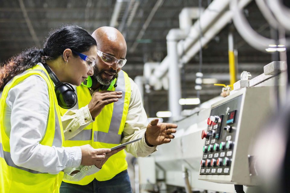 A male and female both in bright yellow safety vests and safety flasses examine the settings on a piece of manufacturing equipment while conducting an industrial energy audit with Ionic Blue LLC