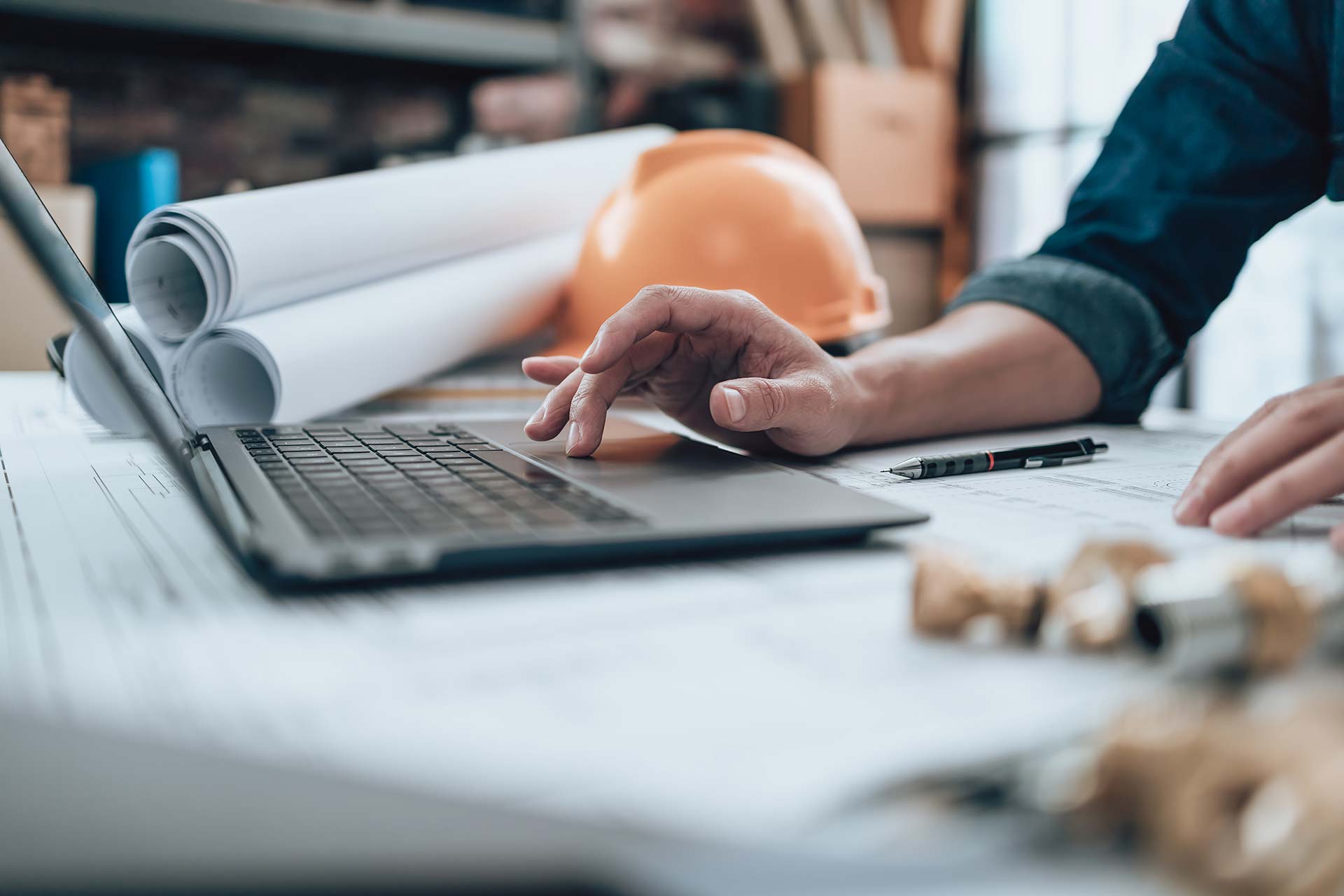 An individuals hands using the mouse pad of a laptop with architectural plans for a facility retrofit with IonicBlue LLC and an orange hard hat in the background